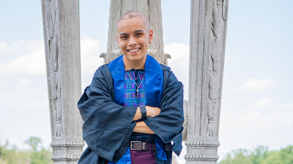 Graduate in academic regalia smiling outdoors in front of stone columns during commencement photoshoot Graduate in academic regalia smiling outdoors in front of stone columns during commencement photoshoot